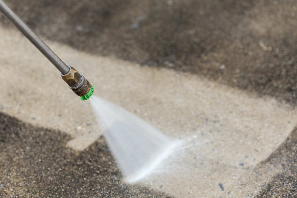 A pressure washer nozzle sprays a powerful stream of water, creating a clean, light-colored path on a dirty, dark gray concrete surface.